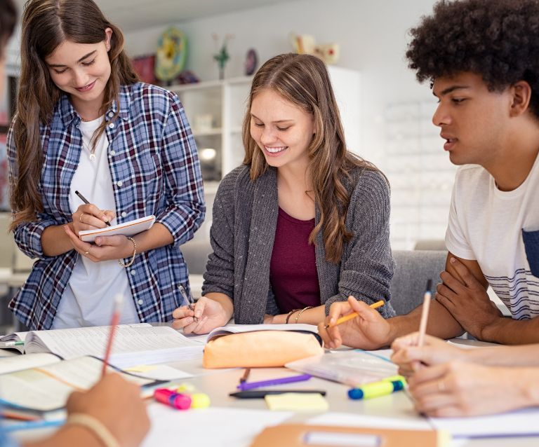 Es sind Schülerinnen und Schüler mit verschiedenen Hautfarben zu sehen, die gemeinsam gut gelaunt an einem Tisch arbeiten. Es ist ein Schulsetting.
