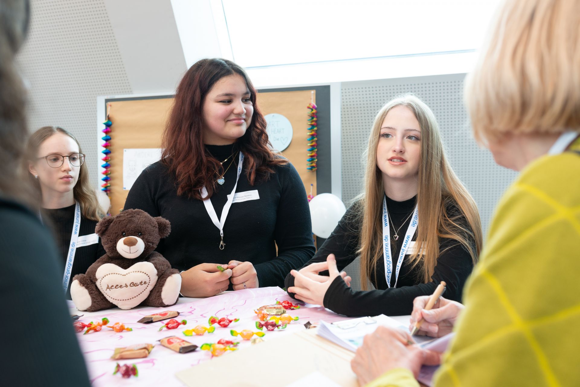Drei Mädchen mit dunklen T-Shirts erzählen von ihrem nachhaltigen Projekt und präsentieren Kuscheltiere. zwei Frauen in gelber und grüner Bluse hören zu.